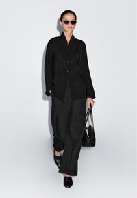 Vrouw die loopt in een zwart oversized blazer en wijde broek, met zwarte zonnebril, een zwarte handtas draagend, tegen een lichte effen achtergrond.