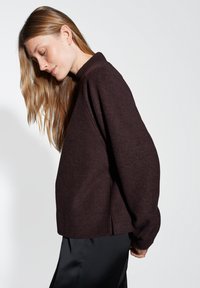Maglione oversize bordeaux in morbido tessuto, caratterizzato da un collo alto e spalle cadenti, con fessure laterali per facilitare il movimento.