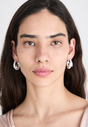 Silver hoop earrings with a textured surface, featuring a twisted design. Model has long dark hair and a natural makeup look.