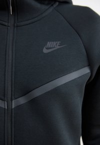 Haut à zip noir en tissu extensible. Présente un logo Nike gris et des accents gris horizontaux sur la poitrine.