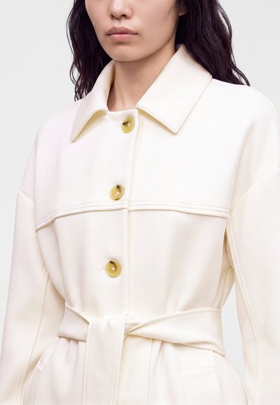 Femme portant un manteau couleur crème avec un large col, trois grands boutons et une ceinture en tissu nouée à la taille.