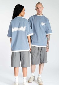 Lyseblå oversized t-shirts med hvid kant og et grafisk design på ryggen, parret med grå shorts og beige sneakers.