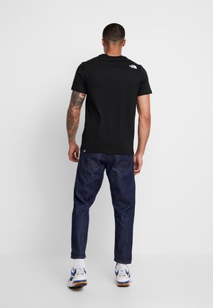 Mann mit tätowiertem Arm, der ein schwarzes North Face T-Shirt, dunkelblaue Jeans und weiße Puma-Sneaker trägt und vor einer schlichten hellgrauen Wand steht.