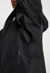 Zwarte lichte jas met een hoge kraag, ritszakken en verstelbaar trekkoord. De stof heeft een gladde, matte textuur.