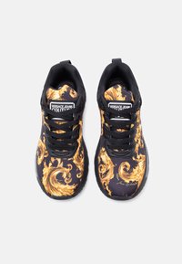 Μαύρα αθλητικά παπούτσια με χρυσό εκκεντρικό σχέδιο, μαύρα κορδόνια και ανάγλυφες λαστιχένιες σόλες. Διαθέτουν λογότυπο στη γλώσσα και χαρακτηριστικά σχεδιαστικά στοιχεία.
