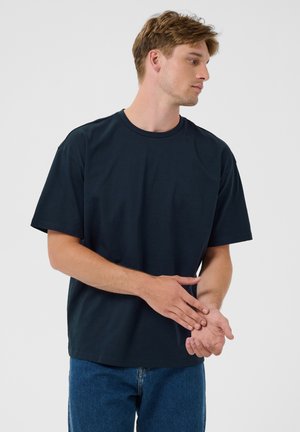 Joven con camiseta azul marino oscuro y jeans azules, mirando hacia un lado, con las manos entrelazadas delante de él sobre un fondo claro y liso.