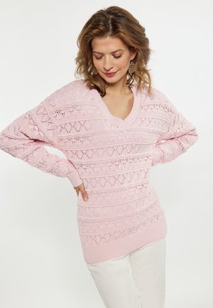 Maglione lavorato a maglia di colore rosa chiaro con scollo a V, caratterizzato da un motivo a diamante in tessuto e polsini a costine, abbinato a pantaloni bianchi.
