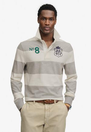 Hombre con camiseta de rugby a rayas beige y gris con el número 8 y escudo, pantalones beige, cinturón trenzado marrón y reloj de pulsera, con las manos en los bolsillos.