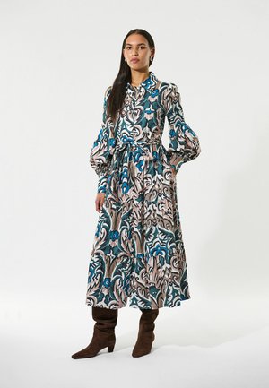Langes Kleid mit tailliertem Oberteil, gepufften Ärmeln und gestuftem Rock. Hat ein florales Muster in Türkis, Rosa und Beige. Wird mit braunen Wildlederstiefeln getragen.