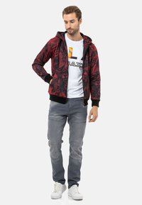 Rote und schwarze gemusterte Kapuzenjacke mit gerippten Bündchen, getragen über einem weißen T-Shirt, kombiniert mit hellgrauen Jeans und weißen Sneakers.