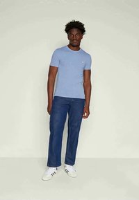 Homem de pele escura a usar uma t-shirt justa azul clara, jeans azul escuro e ténis brancos, posicionado contra um fundo claro e neutro.