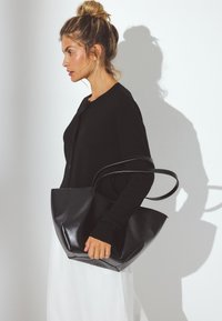 Svart läderhandväska med en strukturerad design och rundade kanter, med två långa handtag. Modellen bär en svart cardigan och en vit kjol.