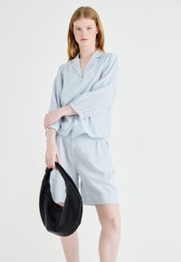 Camicia di lino blu chiaro con chiusura a bottoni e maniche arrotolate, abbinata a pantaloni corti coordinati. Tiene in mano una borsa hobo in pelle nera.