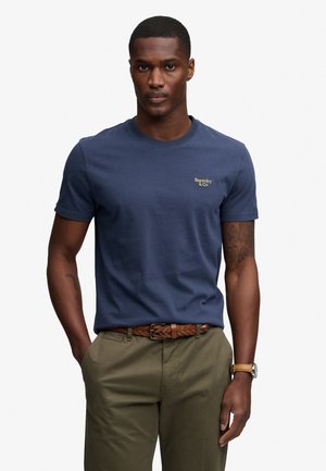 Man die een donkerblauw Superdry T-shirt draagt, olijfgroene broek, gevlochten bruine riem, bruine horloge en een tatoeage op zijn linkerarm toont.