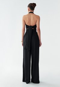 Melna halter top ar atvērtu muguru, sasiets ap jostasvietu. Platas kājas melnas bikses no mīksta auduma. Minimalistisks dizains bez redzamām rakstām.