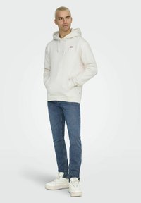 Weißer Hoodie mit Fronttasche und verstellbarem Kordelzug, kombiniert mit blauen Jeans und weißen Sneakern. Einfaches, lässiges Design.