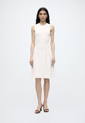 Femme aux longs cheveux foncés portant une robe blanche sans manches jusqu'aux genoux et des talons ouverts noirs, debout devant un fond blanc uni.