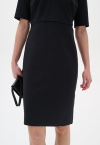 Černé přiléhavé šaty s krátkými rukávy, hladkou texturou a délkou po kolena, doplněné malou černou kabelkou.