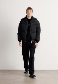 Black puffer jacket with a high collar, zip closure, and quilted texture, worn over a black sweatshirt and pants, completed with black shoes.