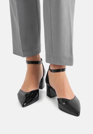 Zapatos de tacón alto de charol negro con punta afilada y correa en el tobillo, con tacón cuadrado y superficie lisa y brillante.