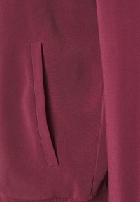 Burgundská mikina s hladkou strukturou, postranní kapsou a žebrovanými manžetami. Materiál vypadá lehký a má neformální design.