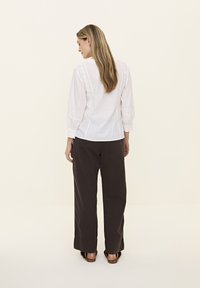 Chemisier blanc à boutons avec dentelle décorative, manches longues bouffantes et ourlet arrondi, associé à un pantalon ample marron foncé.