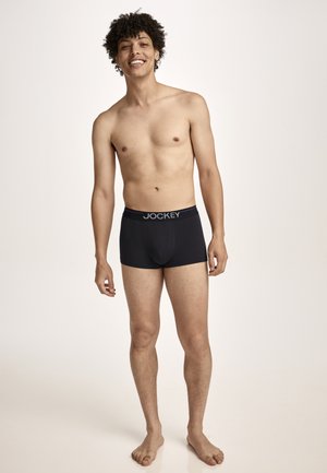 Smiling young man with curly hair wearing black Jockey boxer briefs, standing barefoot against a plain light background.
