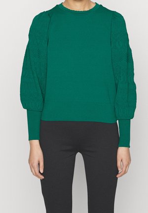 Vrouw draagt een groene top met lange mouwen met gestructureerde pofmouwen en zwarte aansluitende broek tegen een effen achtergrond.