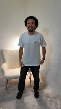 Muž s kučeravými vlasmi oblečený v bielom tričku Nike Athletics a čiernych nohaviciach stojí usmievajúci sa v jemne osvetlenej miestnosti s textúrovaným kreslom a malým stolíkom.