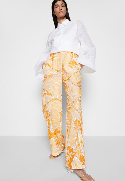 Camisa branca cropped com mangas texturizadas, combinada com calças largas amarelas com um padrão floral branco. Sandálias transparentes completam o look.