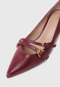 Burgundské kožené lodičky s špičatou špičkou, vybavené dvěma dekorativními pásky a zlatými přezkami přes přední část. Hladká textura s vnitřním podšívkou.