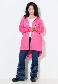 Cardigan con cappuccio rosa chiaro, dotato di tasche anteriori, indossato sopra una maglietta bianca e jeans blu scuro con orli sfrangiati e arruffati. Stivali neri.