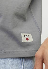 Grijs shirt met korte mouwen van gladde stof, met een witte patch met "R.D.D." en een rode stip. Zichtbare stiksel aan de onderkant.
