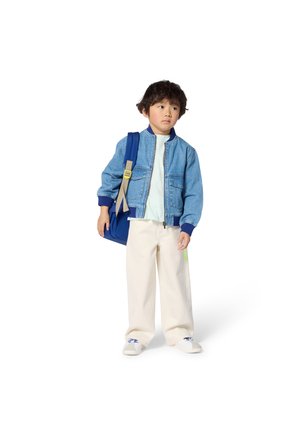 Παιδί που φοράει τζιν μπουφάν bomber, ανοιχτό μπλε μπλουζάκι, παντελόνι φαρδύ εκρού και λευκά αθλητικά παπούτσια, κρατώντας μια μπλε τσάντα ώμου.