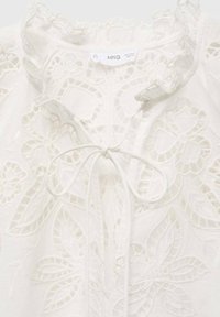 Hvid bluse med blomsterbroderi, en flæset halsudskæring og knytning foran. Tekstureret stof med et delikat blonderdesign.