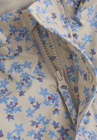 Lätt regnjacka med en krämfärgad bakgrund och blå blommönster. Har tryckknappar och en vertikal dragkedja. Slät textur.