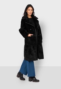 Cappotto di pelliccia sintetica nera con ampio colletto, chiusura doppiopetto e tasche laterali. Abbinato a pantaloni in denim a campana e stivali con tacco a blocco.