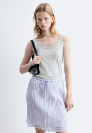 Jeune femme aux cheveux blonds mi-longs porte un débardeur gris clair, une jupe blanche, des créoles et un sac noir à l'épaule.