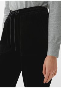 Pantalon en velours côtelé noir avec une texture ribbed, doté d'une taille élastique, d'un cordon de serrage ajustable et de poches latérales. Chemise à manches longues grise visible.