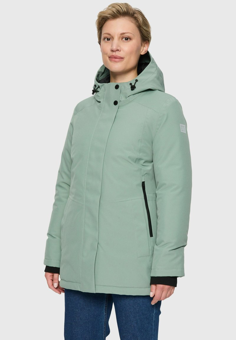 Vrouw in een lichtgroene winterjas met capuchon en zwarte ritszakken, gekleed in donkerblauwe jeans, staande tegen een effen achtergrond.