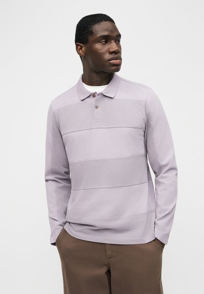 Νέος άνδρας που φοράει ένα μωβ πουκάμισο πόλο με μακριά μανίκια και καφέ παντελόνι, στέκεται με τα χέρια στις τσέπες, κοιτώντας προς τα αριστερά του.