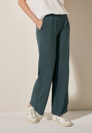 Breedbenige, teal kleurige broek met een elastische tailleband en zijzakken, gecombineerd met witte sneakers met groene accenten. Glad textuur van de stof.