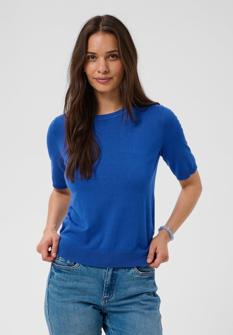 Femme aux longs cheveux bruns portant un pull bleu vif à manches courtes et un jean bleu, posant avec les mains près des poches.