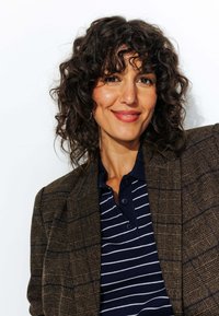 Blazer écossais marron sur un polo rayé bleu marine. La texture est tissée, avec une finition polie. Cheveux bouclés et foncés. Fond blanc.