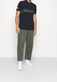 Mörkblå polo skjorta med grön horisontell rand som har "HACKETT"-logotypen i vitt, kombinerad med olivgröna cargobyxor och vita sneakers.