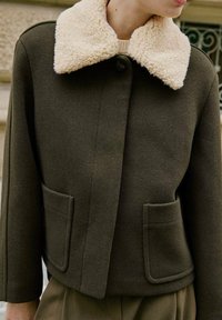 Chaqueta verde oscuro con un cuello de borreguillo crema, que cuenta con dos bolsillos frontales y un cierre de botones. Tejido texturizado con un ajuste estructurado.
