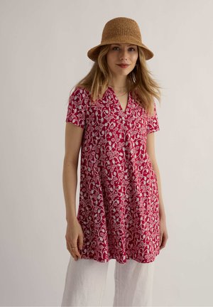 Žena oblečená v červenom kvetinovom tunike, bielych nohaviciach, bledohnedom tkanom klobúku a s minimom šperkov, stojaca pred jednoduchým svetlým pozadím.