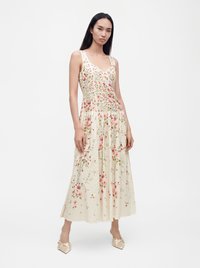 Kvinde stående iført en ærmeløs cremefarvet kjole med pink blomsterprint og beige spidse flade sko mod en ensartet hvid baggrund.