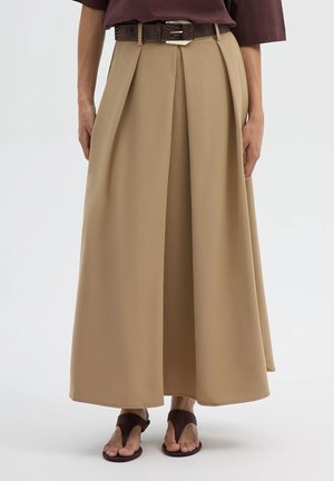Donna che indossa una gonna lunga beige con piega centrale, cintura marrone e sandali marrone scuro, in piedi su uno sfondo semplice.