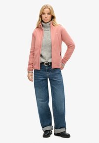Puszysta różowa polarowa kurtka z wysokim kołnierzem, zestawiona z szarym swetrem na stojąco, luźnymi niebieskimi dżinsami i czarnymi butami. Prosty, casualowy design.